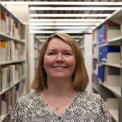 A woman with shoulder-length blonde hair smiles in a library, wearing a black and white patterned blouse.